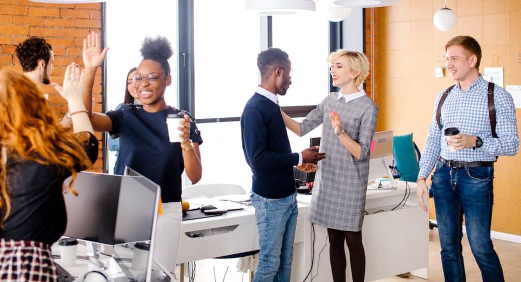 two diverse women at recruiting event are making high five, Caucasian man is looking at talking woman with fair hair with African man in break time